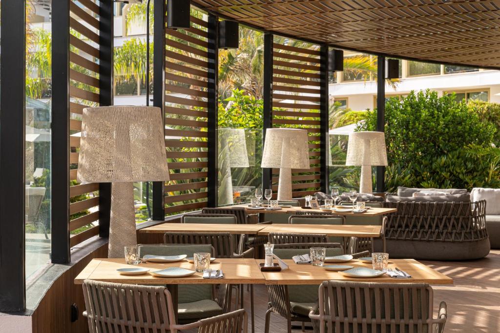 Mūsdienīgs restorāns ar koka mēbelēm un lieliem logiem, caur kuriem redzams zaļš dārzs. Interjers ir elegants un gaisīgs, radot patīkamu atmosfēru.