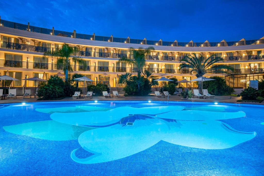 Mūsdienīgs viesnīcas komplekss ar zilu baseinu, kura centrā ir zieda motīvs, un apgaismotām terasēm, kas rada relaksējošu atmosfēru tumšajā vakarā.