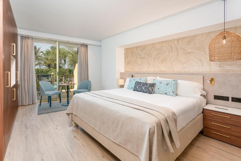 Mūsdienīga viesnīcas numura iekšskats ar lielu gultu, dekoratīviem spilveniem un skatu uz palmu dārzu caur stikla durvīm. Numurā ir ērti krēsli un gaismas apgaismojums, radot relaksējošu atmosfēru.