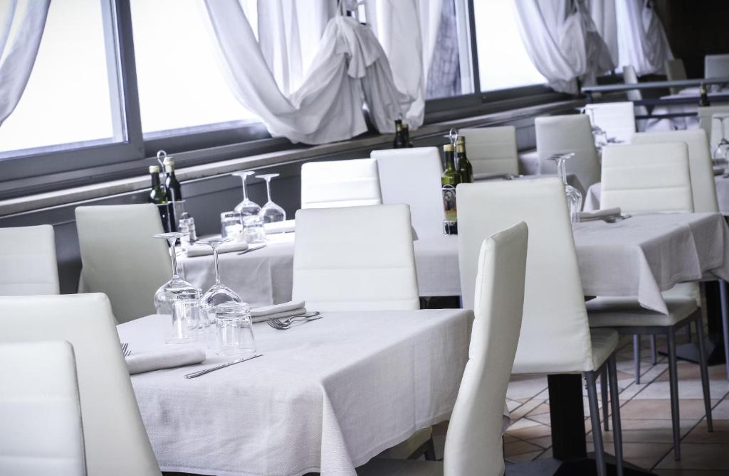 Mūsdienīgs restorāna interjers ar baltām galdautām un elegantiem galdiem, kas sagatavoti viesu uzņemšanai. Langu apstādījumi rada gaisīgu un svaigu atmosfēru.