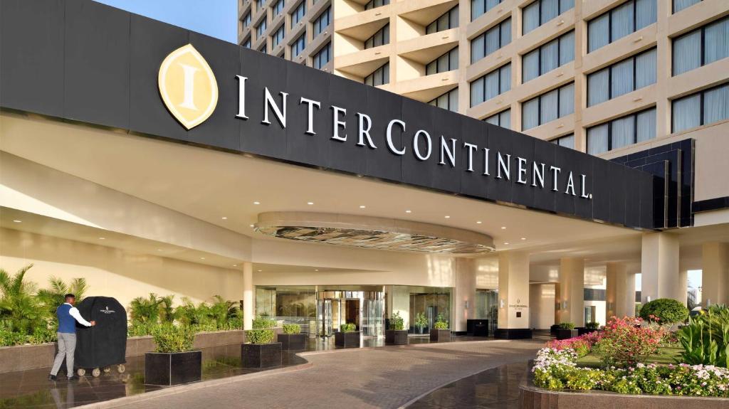 InterContinental viesnīcas ieeja ar ziedu kompozīcijām un viesmīli, kas stumj bagāžu. Būvēta moderna arhitektūra un viesmīlīga atmosfēra.