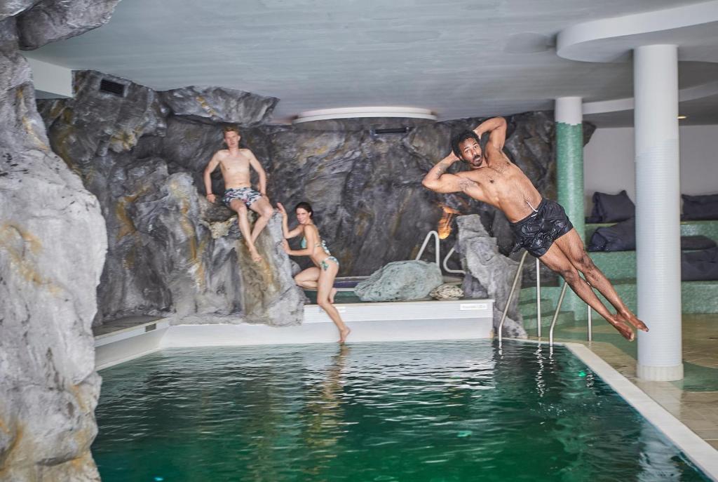 Trīs cilvēki, divi sēžot un viens lēkājot, izklaidējas baseinā, kas iekārtots dabas stila vidē. Ūdens ir dzidrs un apkārt ir akmeņi un ērti sēdēšanas krēsli.