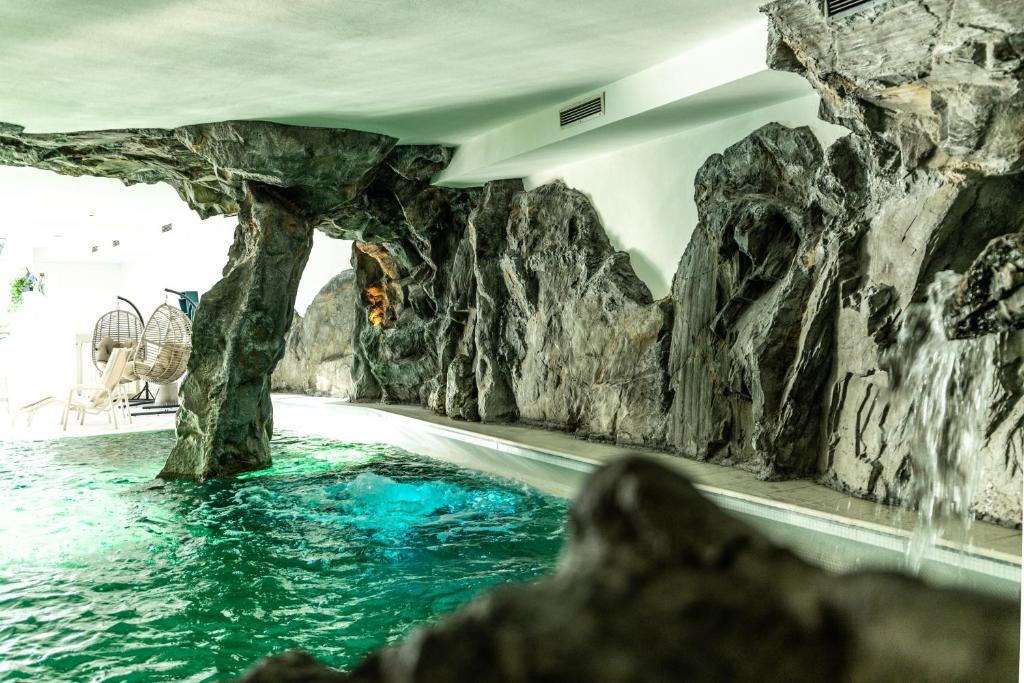 Iekštelpu baseins ar akmens struktūrām un zaļganu ūdeni, radot relaksējošu atmosfēru. Redzami arī komfortabli krēsli un ventilatori.