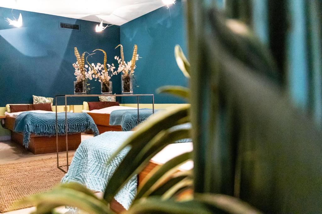 Mūsdienīgs relaksācijas telpas interjers ar zilu sienu, divām gultām un dekoratīvām puķēm. Telpa ir apgaismota ar gaismekļiem, radot mierīgu atmosfēru.