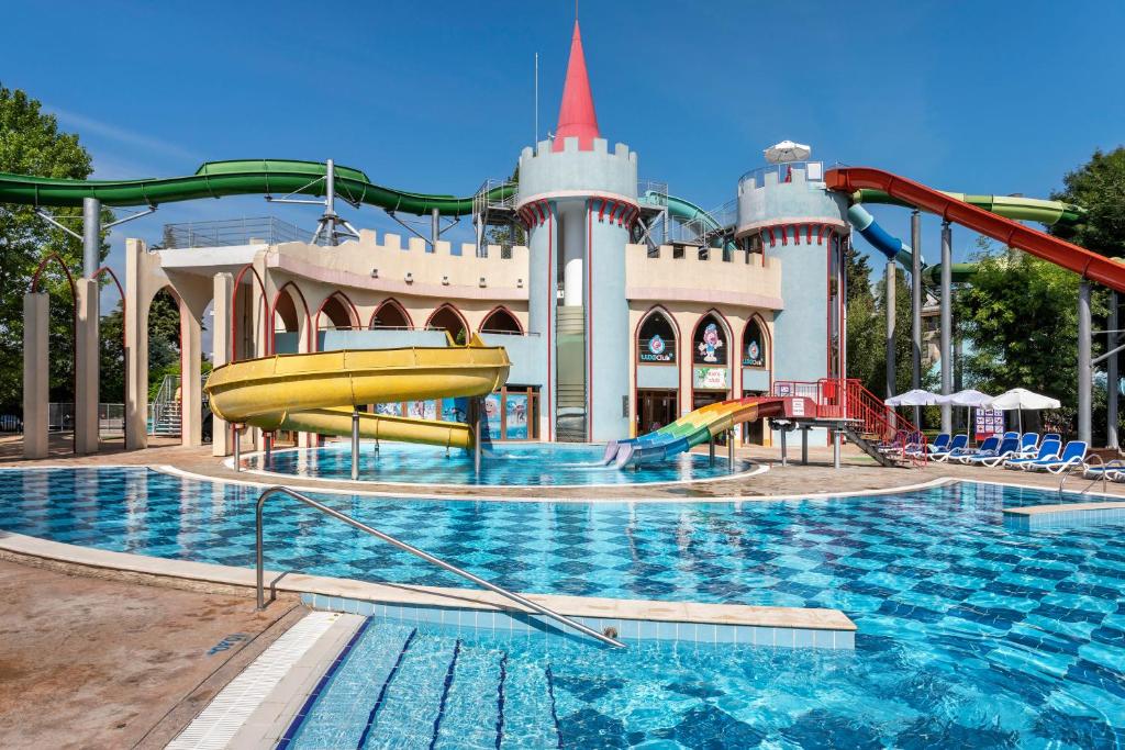 Ūdens atrakciju parks ar krāsainiem ūdens slidkalniņiem un zilu baseinu, kas atrodas saulainā dienā. Apskatāma viduslaiku stila ēka ar torņiem un slīdošām struktūrām.