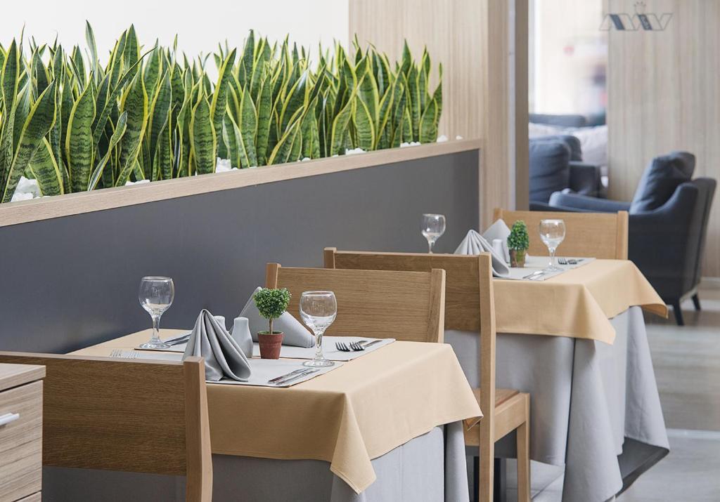 Mūsdienīgs restorāna interjers ar koka mēbelēm un dabiskiem augiem, kas rada patīkamu atmosfēru. Galdi ir klāti ar dzelteniem galdautiem, un uz tiem ir glāzes un galda piederumi.