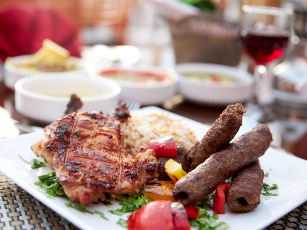 Svaigi pagatavota maltīte, kas ietver grilētu vistu, kebabus, rīsus un dārzeņu garnējumu, uz baltas šķīvja. Fons ar krāsainām zupām un sarkano vīnu.