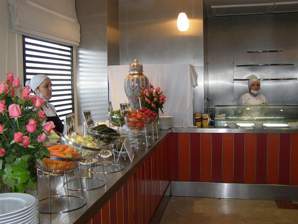 Buffet stila galdi ar svaigiem dārzeņiem un augļiem, ko pasniedz restorāna darbinieki. Interjerā redzami rozā rozes un elegants dizains.