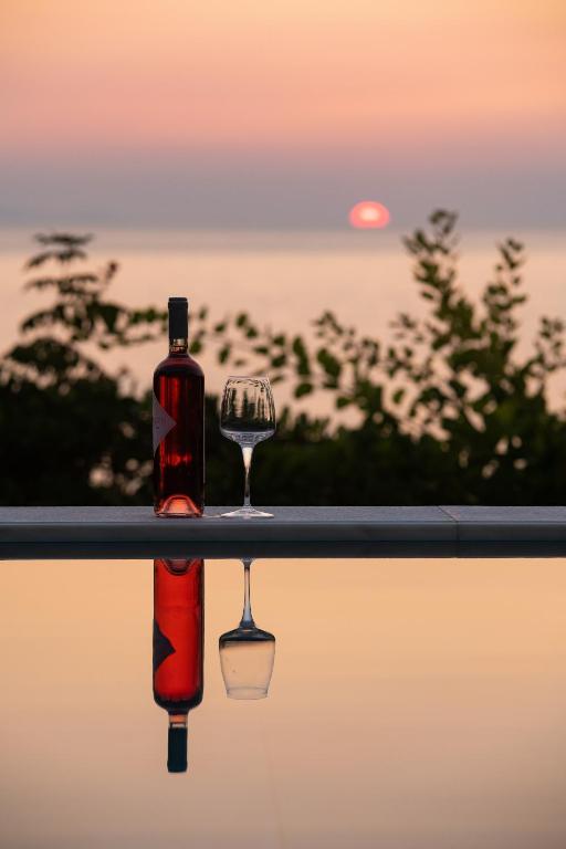 Rozā vīna pudele un glāze stāv pie baseina, atspoguļojot saulrietu un mierīgu jūras ainavu.