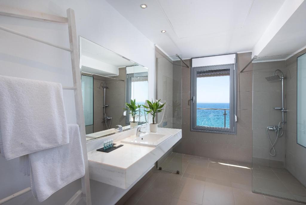 Mūsdienīgs vannas istabas interjers ar skatu uz jūru, baltiem dvieļiem un elegantu izlietni. Gaismas un telpas harmonija rada relaksējošu atmosfēru.