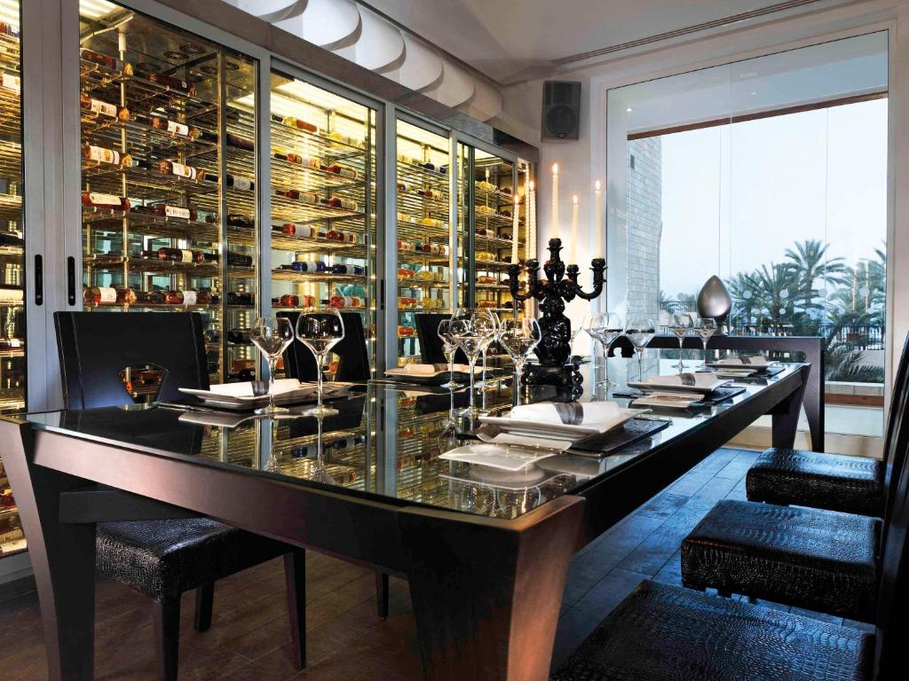 Mūsdienīga ēdamzāle ar elegantu galdu, stikla vāku un melnu svečturi, fonā redzama vīnu glabātuve ar dažādām pudelēm. Interjers ir gaišs un gaumīgs, radot patīkamu atmosfēru.