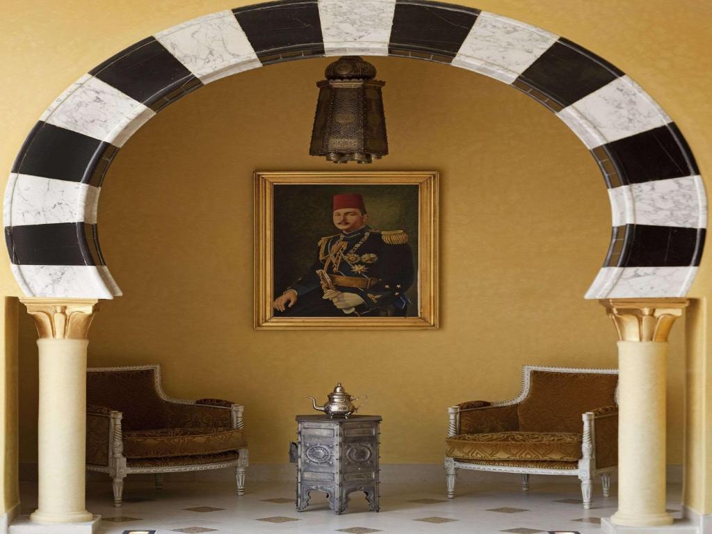 Elegantā interjera skats ar arku, kas izrotāta ar melnbaltām flīzēm un lampu, blakus atrodas divas greznas krēsli un sudraba tējkanna uz maza galda. Fons rotā portrets, kas ievērojamā veidā papildina telpas atmosfēru.