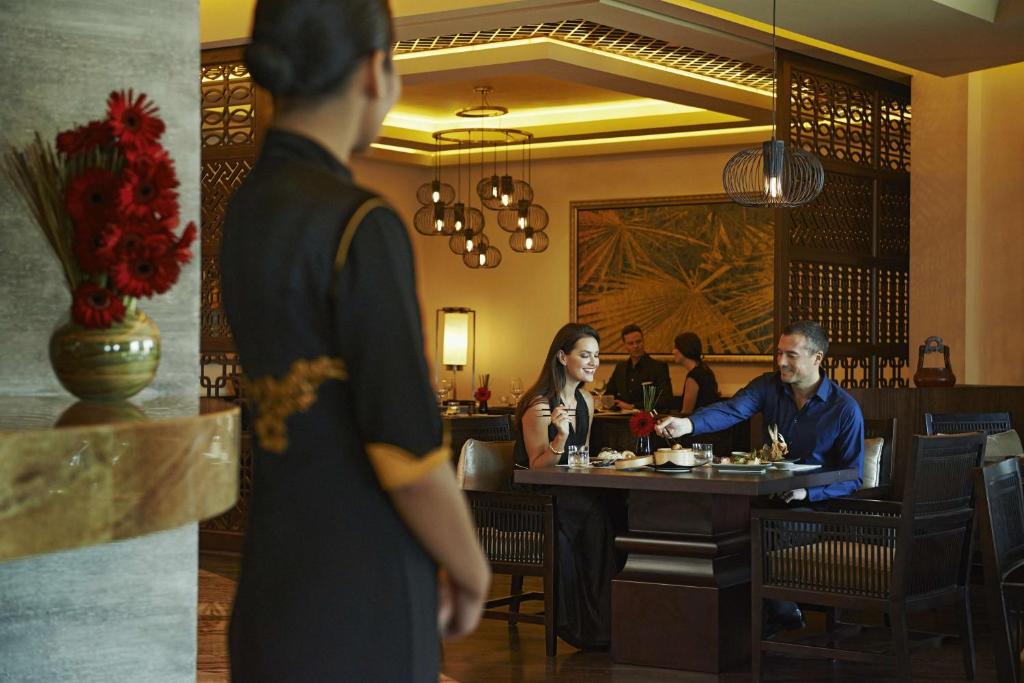 Eleganta restorāna atmosfēra, kur pāris baudījuši maltīti, un viesmīle apkalpo. Siltas gaismas un moderns interjers rada patīkamu noskaņu.