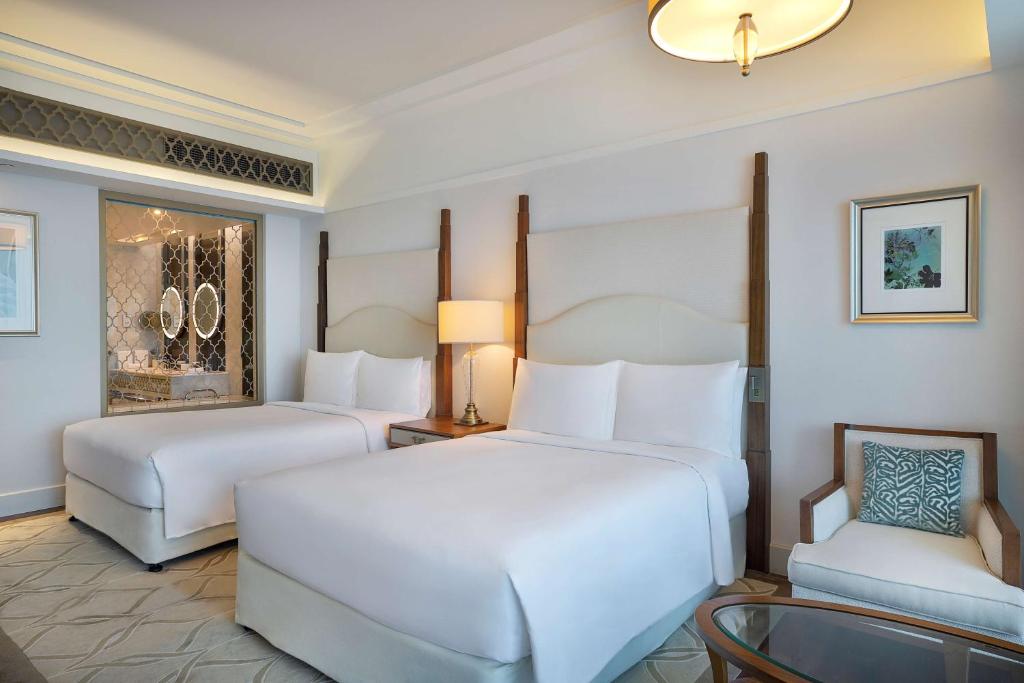 Moderns viesnīcas numurs ar divām gultām, gaišām sienām un elegantu interjeru, kas ietver atpūtas krēslu un lampu. Fons ir redzams vannas istabas skats ar spoguļiem.