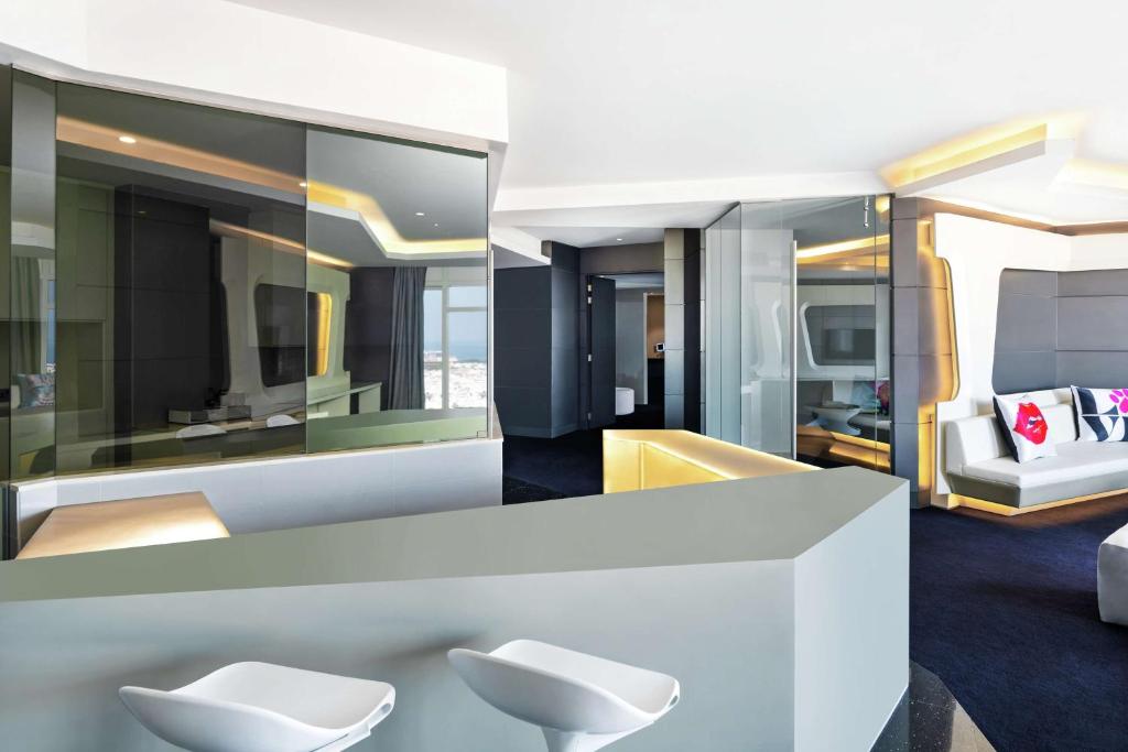 Mūsdienīga viesnīcas istaba ar stikla sienām, elegantām mēbelēm un izsmalcinātu apgaismojumu. Telpa ir plaša un gaiša, ar skatu uz pilsētu.