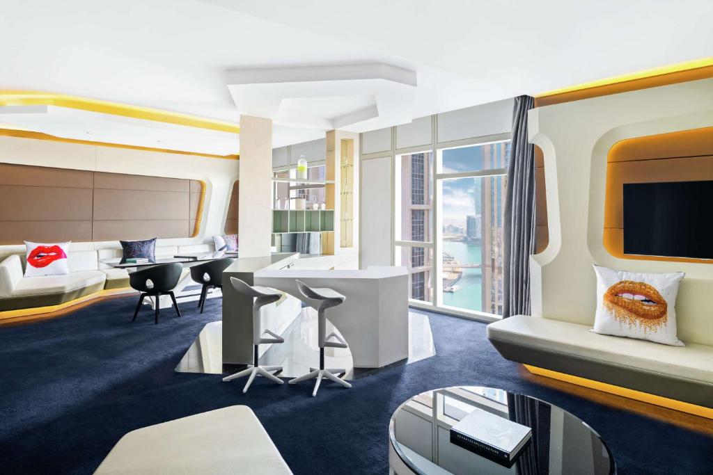 Mūsdienīgs viesnīcas numurs ar stilīgu interjeru, kas ietver komfortablu dīvānu, elegantus krēslus un lielus logus ar skatu uz pilsētu. Numurā ir arī moderns galdiņš un spilveni ar mākslinieciskām motīvām.