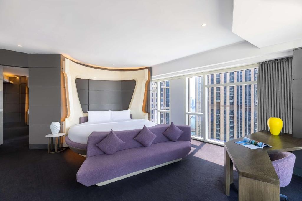 Moderns viesnīcas numurs ar apaļu gultu un stilīgu interjeru, kas ietver purpura krāsas mēbeles un lielus logus ar skatu uz pilsētu. Numurā ir arī darba galds un dekoratīva vāze.