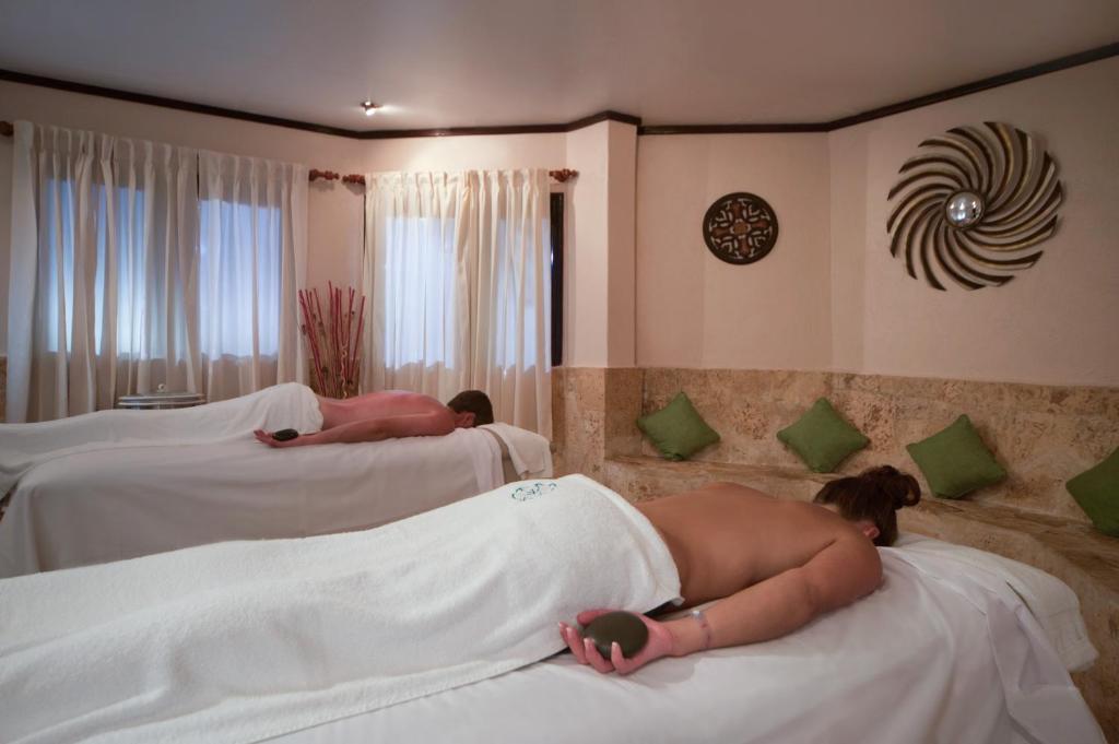 Relaksējoša spa sesija diviem, kur abi apmeklētāji guļ uz masāžas galdiem, pārklāti ar baltām dvielīšiem, kamēr apkārt ir mierīga un estētiska vide.