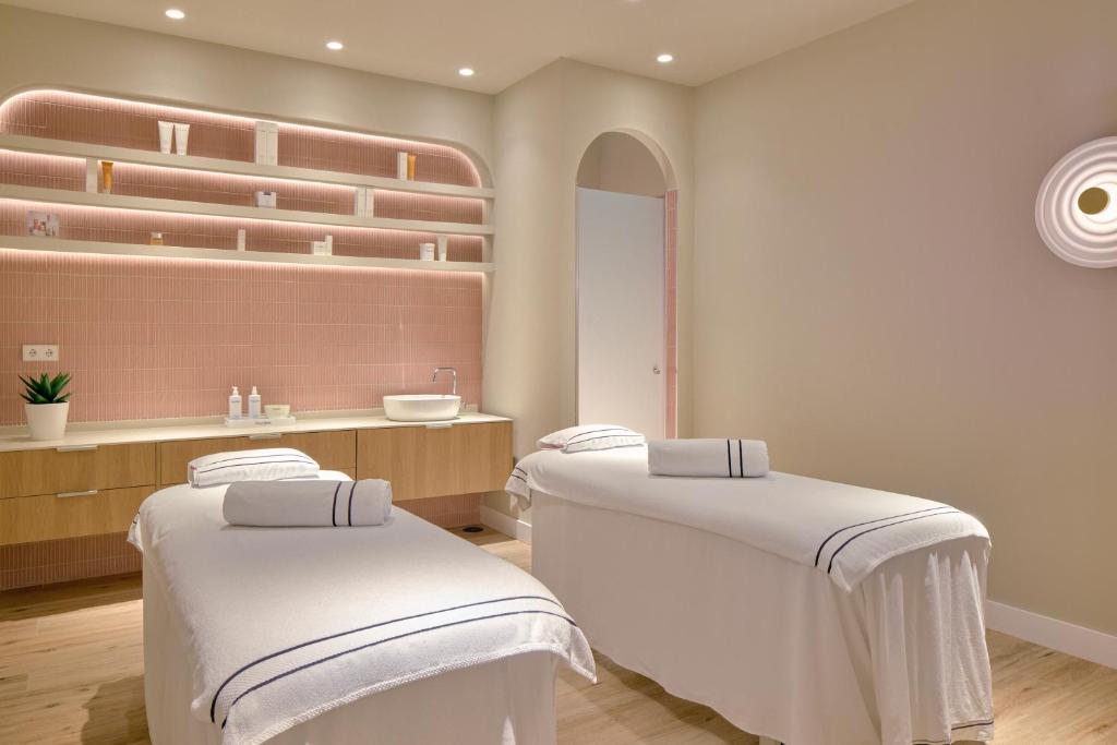 Mūsdienīgs spa telpa ar divām masāžas galdiem, kas pārklāti ar baltām dvielēm. Aiz galdiem redzama gaiši rozā sienas apdare ar plauktiem, kuros izvietoti kosmētikas līdzekļi un augs. Apgaismojums ir maigs un relaksējošs.