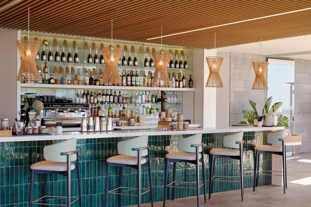 Mūsdienīgs bārs ar elegantu dizainu, kas ietver rattan lampas un plašu alkohola izvēli. Bāra letes priekšplānā ir stilīgas krēslas un zaļš flīžu apdare.
