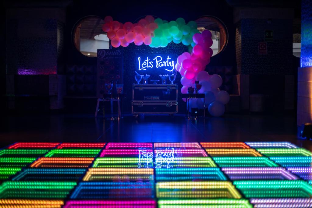 Krāsaina deju grīda ar gaismas efektiem un "Let's Party" neona zīmi, fonā redzami baloni un galdiņš ar dzērieniem.
