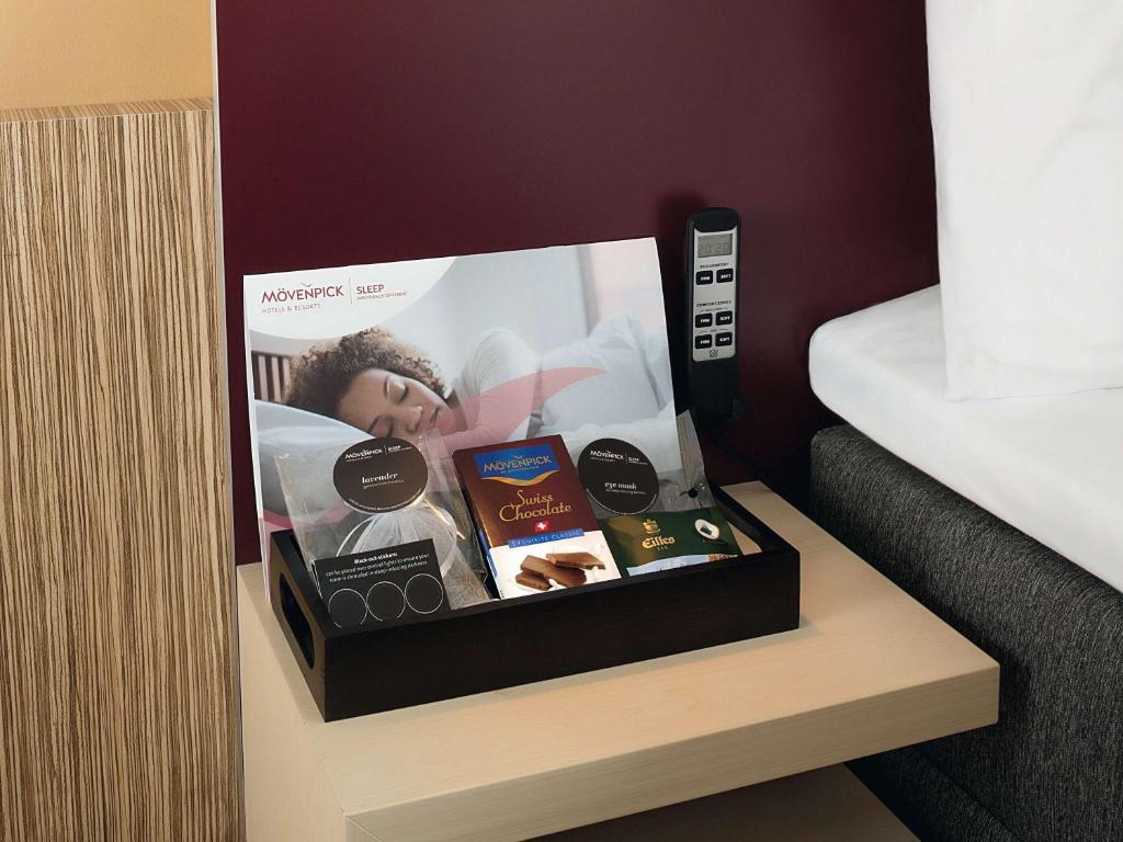 Mēbeļu tray ar Movenpick Sleep komplektu, kas ietver relaksējošas preces, piemēram, acu masku un šveiciešu šokolādi, atrodas uz naktsgaldiņa viesnīcas numurā.