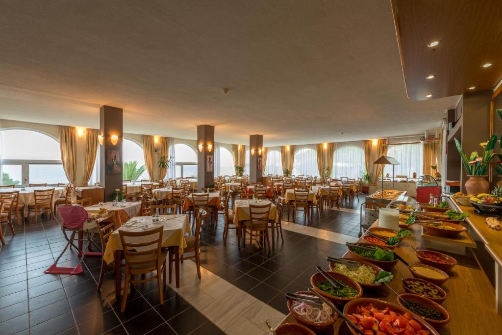 Gaiša restorāna iekārtojums ar plašiem logiem, kas piedāvā skatu uz jūru, un bagātīgu ēdienu izvēli uz bufetes galda. Eleganti galdi un krēsli rada patīkamu atmosfēru maltītēm.