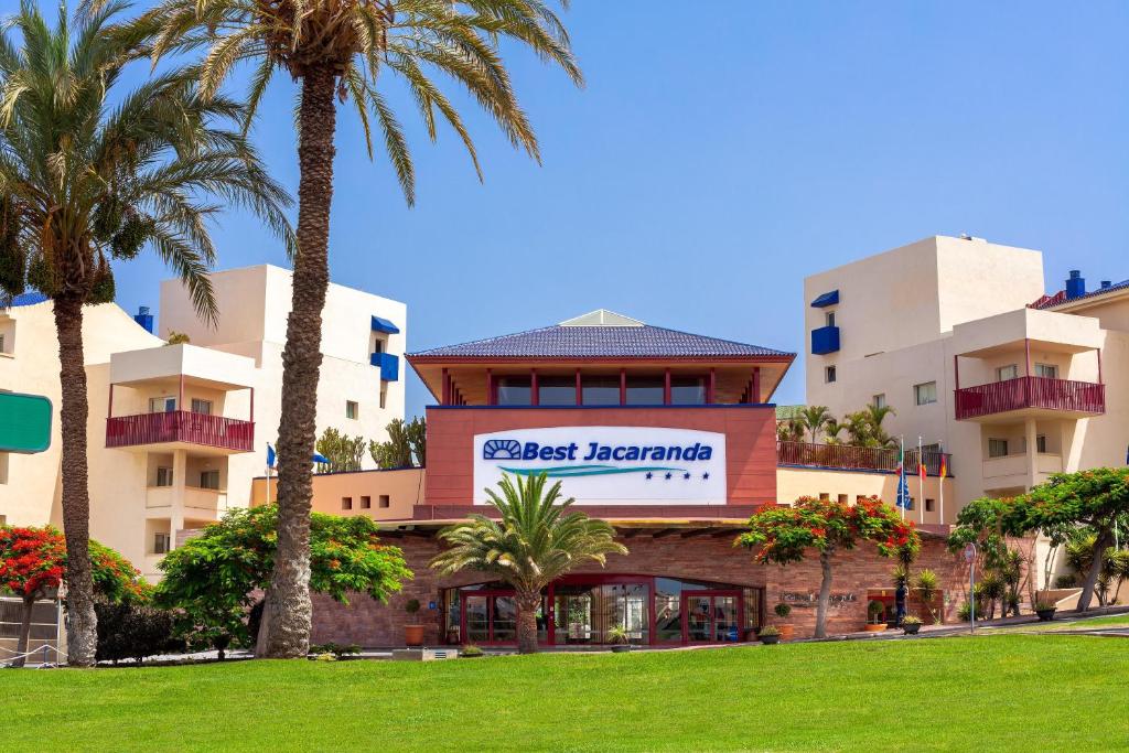 Viesnīcas Best Jacaranda ieeja ar palmām un ziedošiem krūmiem apkārtnē, skaidrā debesīs.