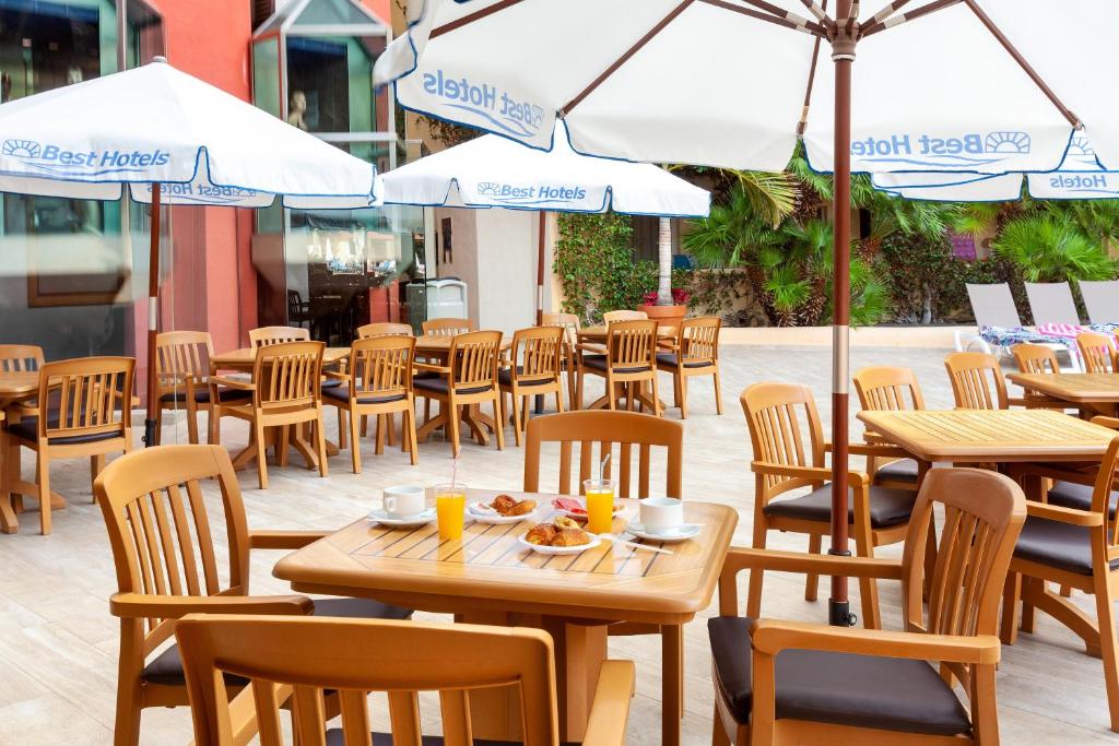 Āra kafejnīca ar koka galdiem un krēsliem, kas apgādāti ar saulessargiem. Uz galda ir pasniegti brokastu ēdieni un dzērieni, radot patīkamu un relaksējošu atmosfēru.