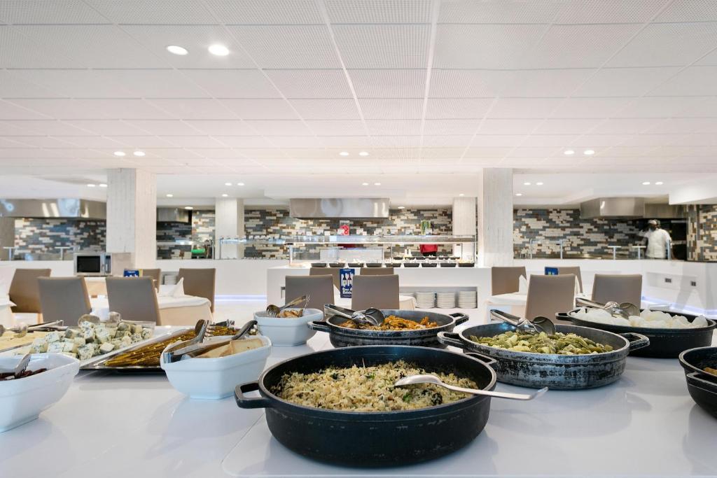 Moderns bistro ar dažādiem ēdieniem, kas izkārtoti uz galda. Fons ietver atvērtu virtuvi ar stilīgām flīzēm un ērtām ēdināšanas vietām.