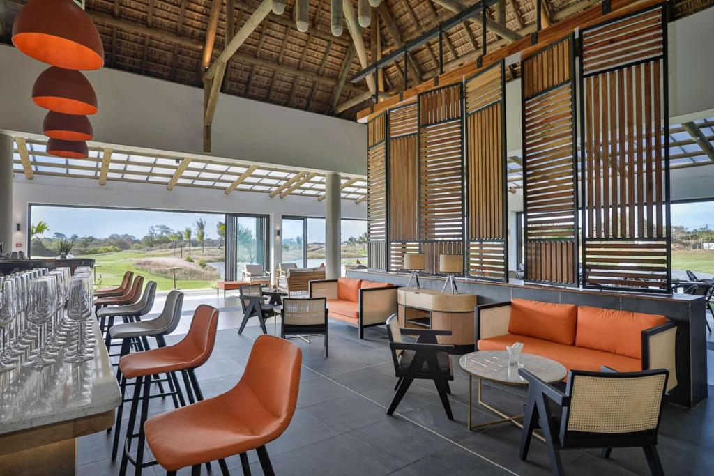 Mūsdienīgs bārs ar stilīgu interjeru un plašiem logiem, kas piedāvā skatu uz ainavu. Sēdvietas ar oranžiem spilveniem un elegantu galdu veido relaksējošu atmosfēru.