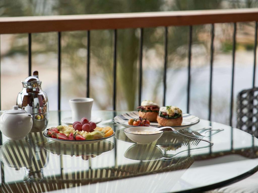 Broka stalā ir dažādi augļi, kafija un maizītes ar dārzeņiem, kas novietoti uz stikla galda uz balkona.