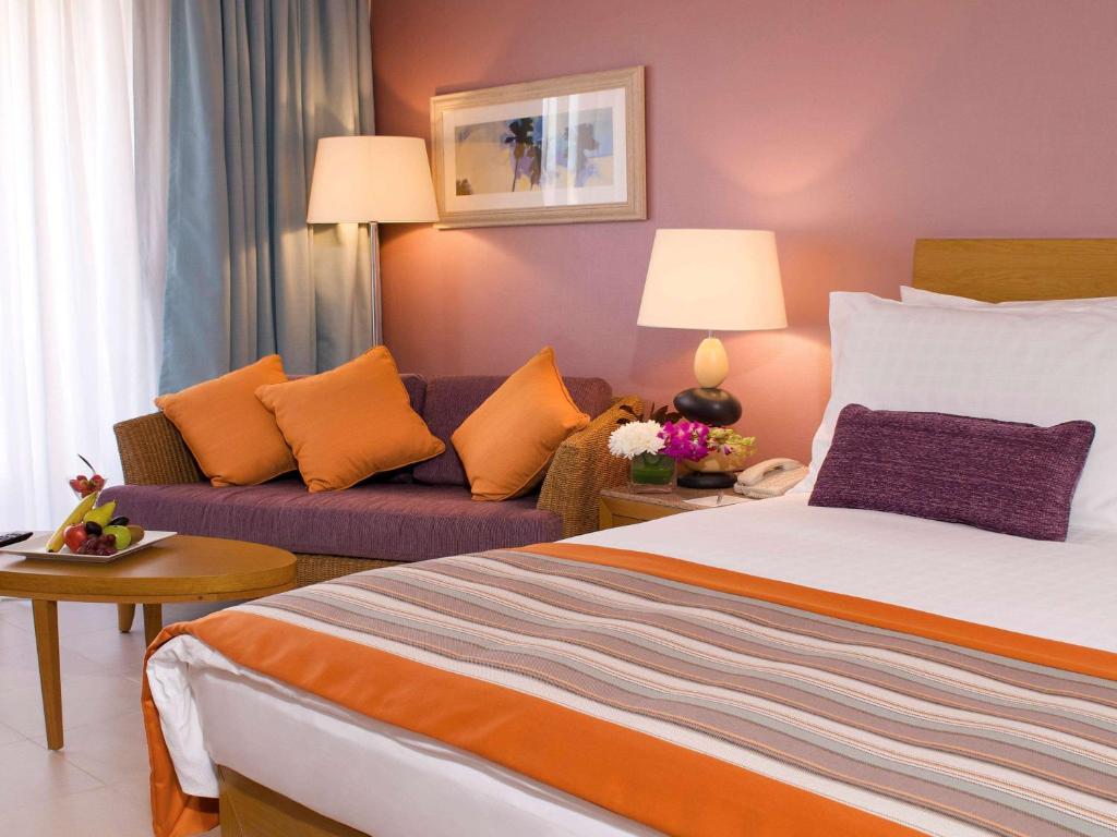 Mūsdienīga viesnīcas istaba ar ērtu gultu, dīvānu un dekoratīvām spilveniem, pie galda ar augļiem. Sienas rotā mākslas darbs, un telpā ir silta apgaismojuma atmosfēra.