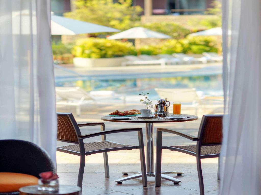 Rāmja skats uz galdu ar brokastīm, kafiju un sulu, kas novietots pie baseina, apstādījumos un sauļošanās krēslos.