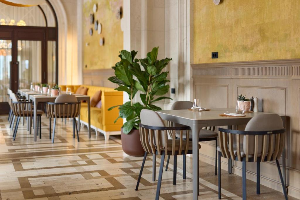 Mūsdienīgs restorāna interjers ar dzeltenu dīvānu, elegantiem galdiem un krēsliem, zaļām augu kompozīcijām un māksliniecisku sienas apdari.