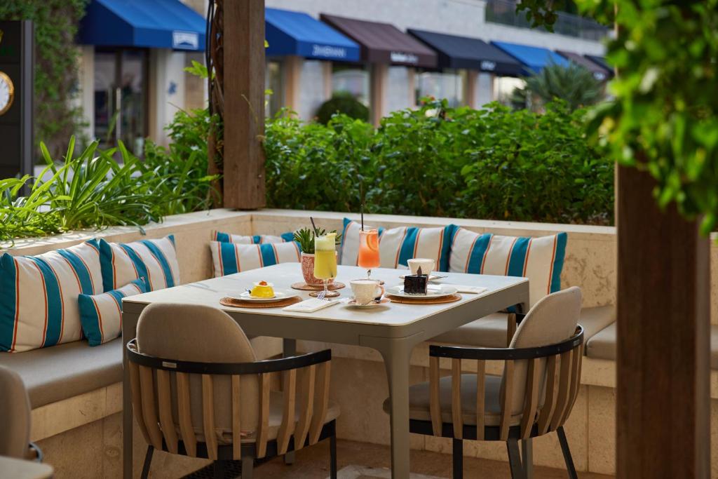 Eleganta terase ar komfortablām mēbelēm, kur uz galda novietoti dzērieni un deserti. Apkārt augu zaļums un krāsainas spilvenu detaļas.
