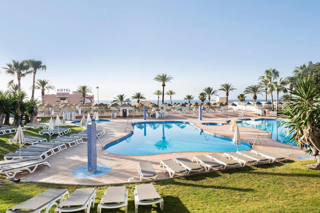 Viesnīcas baseins ar palmām un sauļošanās krēsliem, skats uz jūru un saulainu debesi. Ideāla vieta atpūtai un relaksācijai.