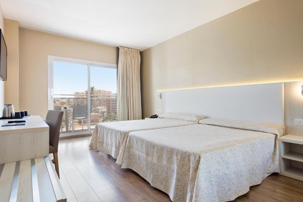 Mūsdienīga viesnīcas istaba ar divām gultām, galdu un skatu uz pilsētu caur lielajiem logiem. Iekārtojums ir gaišs un elegants, radot komfortablu atmosfēru.