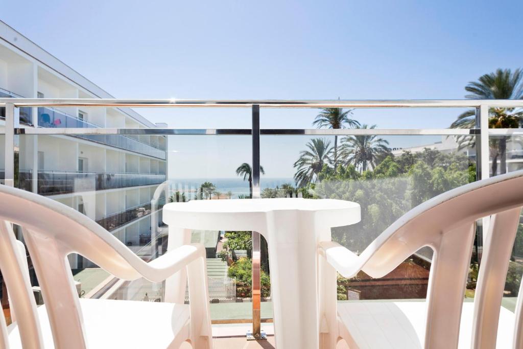 Balkons ar skatu uz jūru, kurā redzami divi baltie plastmasas krēsli un galds, apkārt aug palmām un zaļumiem. Ideāla vieta relaksācijai un atpūtai.