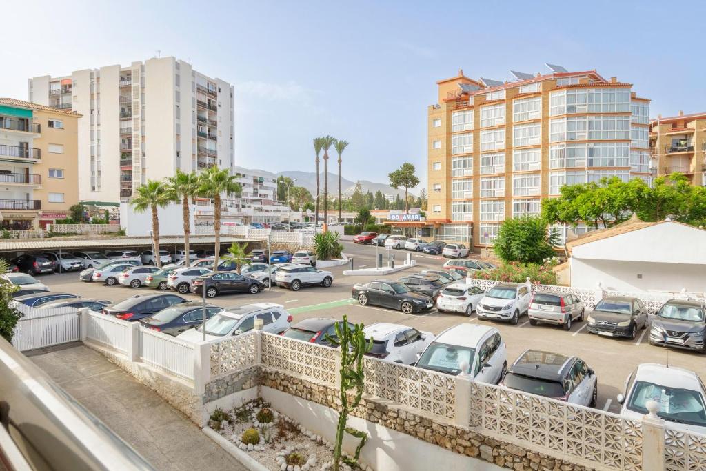 Skats uz pilsētas ainavu ar daudzdzīvokļu mājām, kokiem un automašīnām stāvlaukumā. Apkārtne ir apgaismota, ar saulainu laiku un kalnu fona.