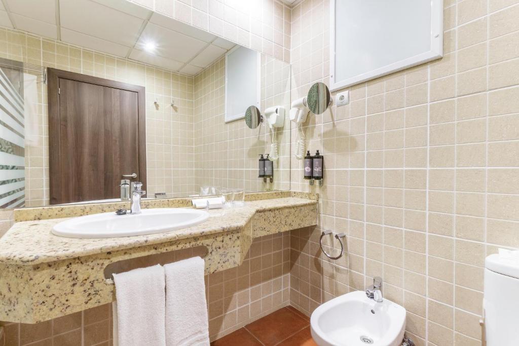 Moderns vannas istabas interjers ar granīta izlietni, spoguli un higiēnas piederumiem uz sienas. Siltas krāsas flīzes un koka durvis rada mājīgu atmosfēru.