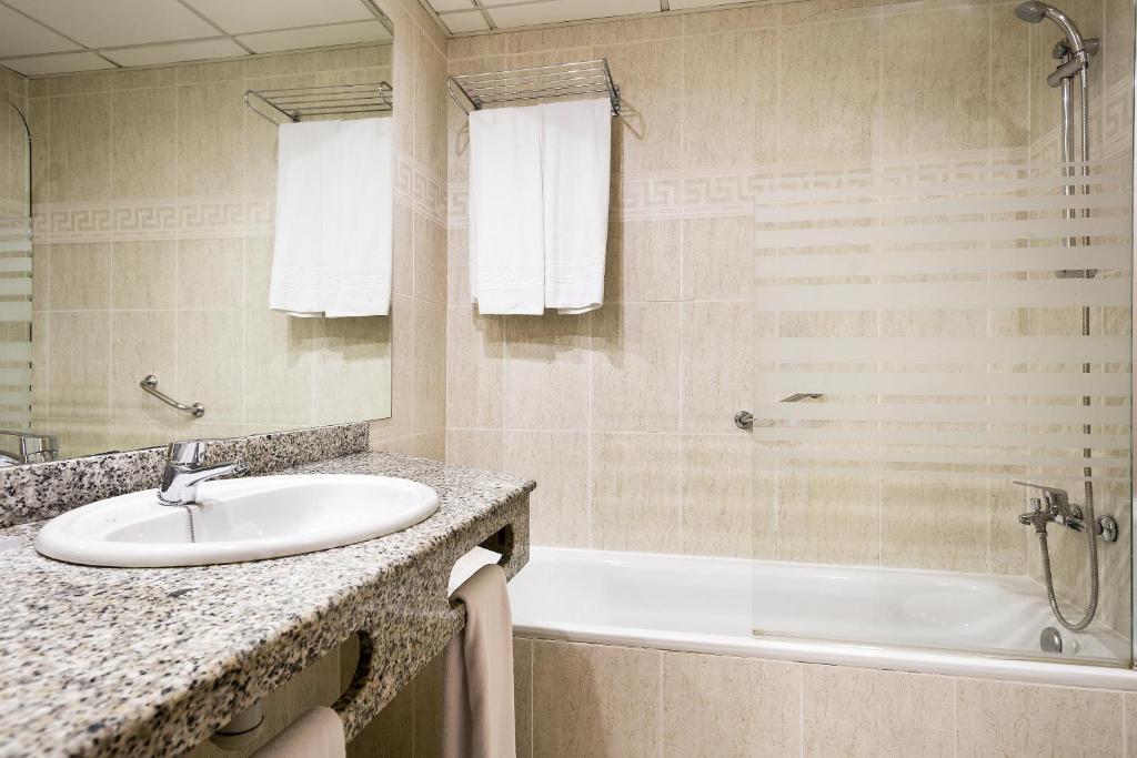 Mūsdienīga vannas istaba ar akmens virsmu, vannu un spoguli. Svaigi baltas dvieļi un stilizēts sienu dizains papildina telpas estētiku.