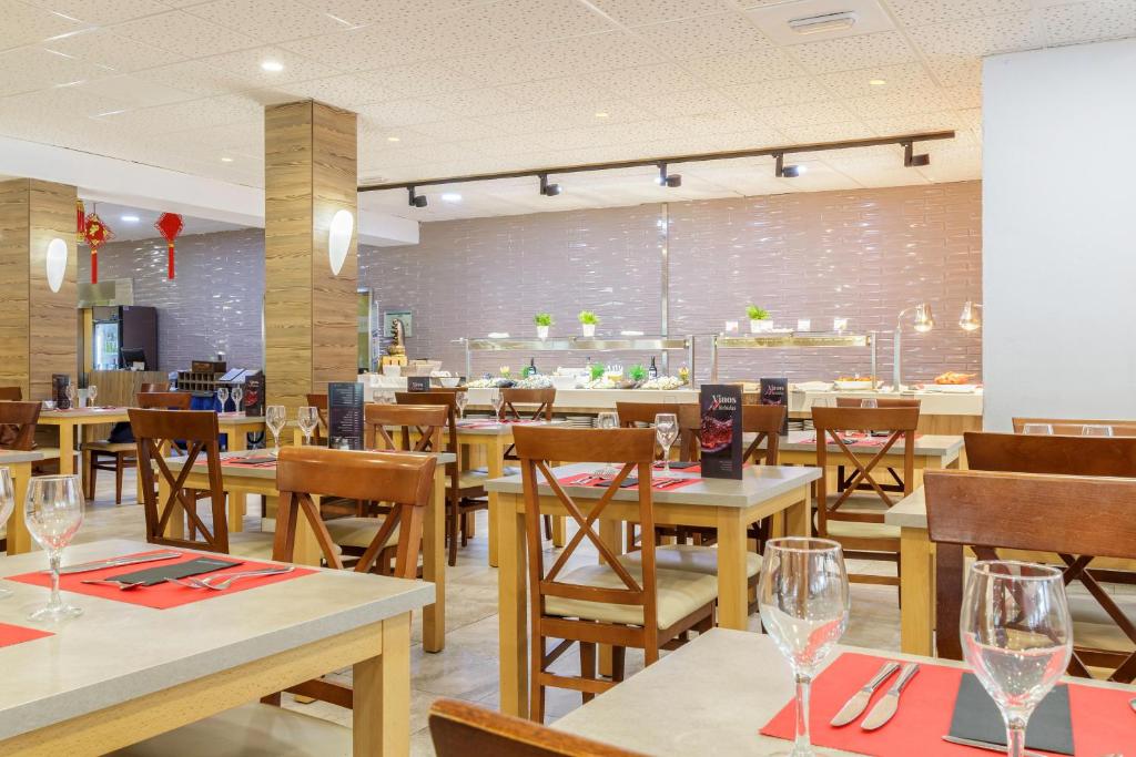 Mūsdienīgs restorāns ar koka mēbelēm un elegantu galdu iestatījumu, kurā redzama bufete ar dažādiem ēdieniem un zaļajiem dekoratīvajiem augiem.