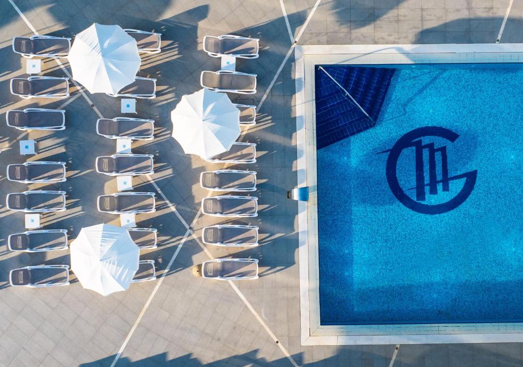 Peldbaseins ar zilu ūdeni un stilīgu logotipu, apkārt novietotas sauļošanās krēsli un saulessargi. Ideāla vieta atpūtai un relaksācijai.