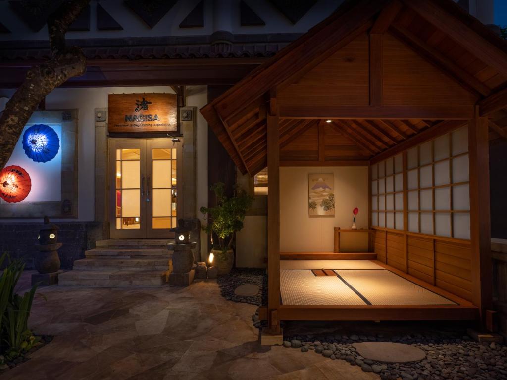 Mūsdienīga japāņu stila ēka ar koka fasādi, izgaismota ieeja un tradicionāls tatami grīdas laukums. Apgaismojums izceļ krāsainus papīra lietussargus un akmens lampas, radot mierīgu atmosfēru.