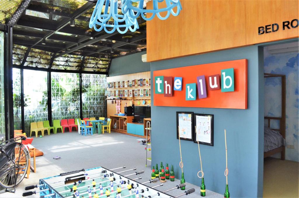 Radošs bērnu klubs ar krāsainām mēbelēm, foosball galdu un interaktīvām aktivitātēm, kas veidots gaišā un atvērtā vidē. Telpā ir redzamas dažādas rotaļlietas un uz sienām izvietoti izglītojoši materiāli.