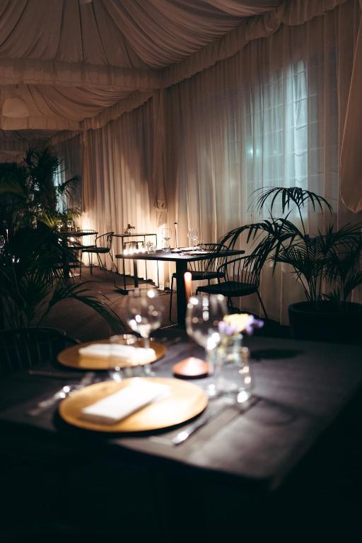 Elegants restorāna interjers ar romantisku apgaismojumu, ko veido ziedi un sveces. Melnā galdautā novietoti zelta šķīvji un glāzes, radot intīmu atmosfēru.