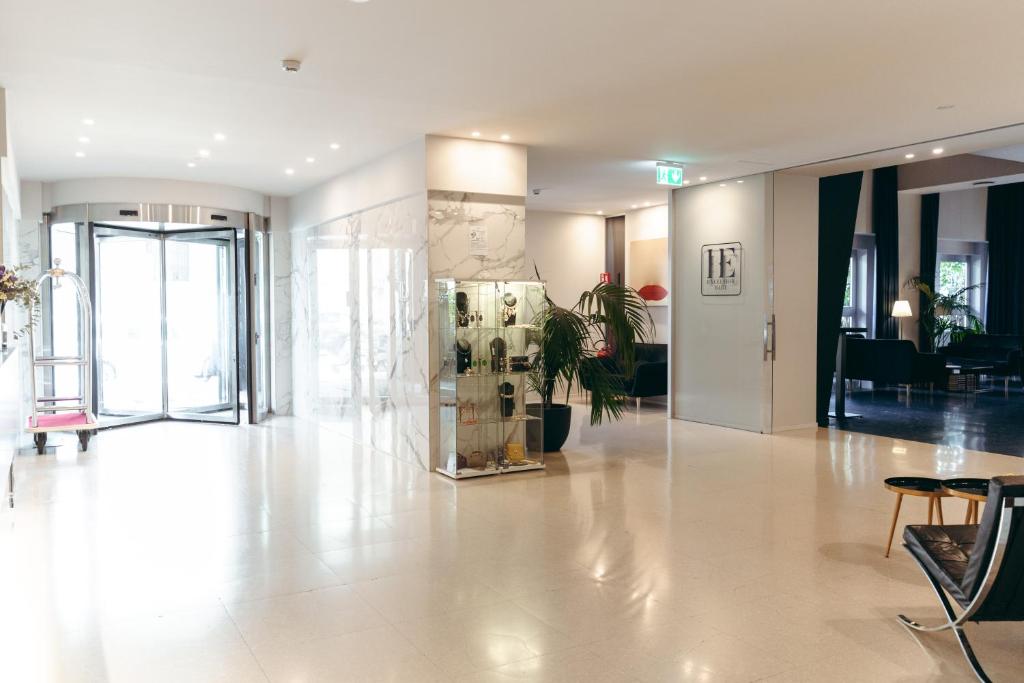 Mūsdienīgs viesnīcas vestibils ar rotējošām durvīm, stilīgu stikla vitrīnu un augstām augu dekorācijām. Telpa ir gaiša un plaša, radot viesmīlīgu atmosfēru.