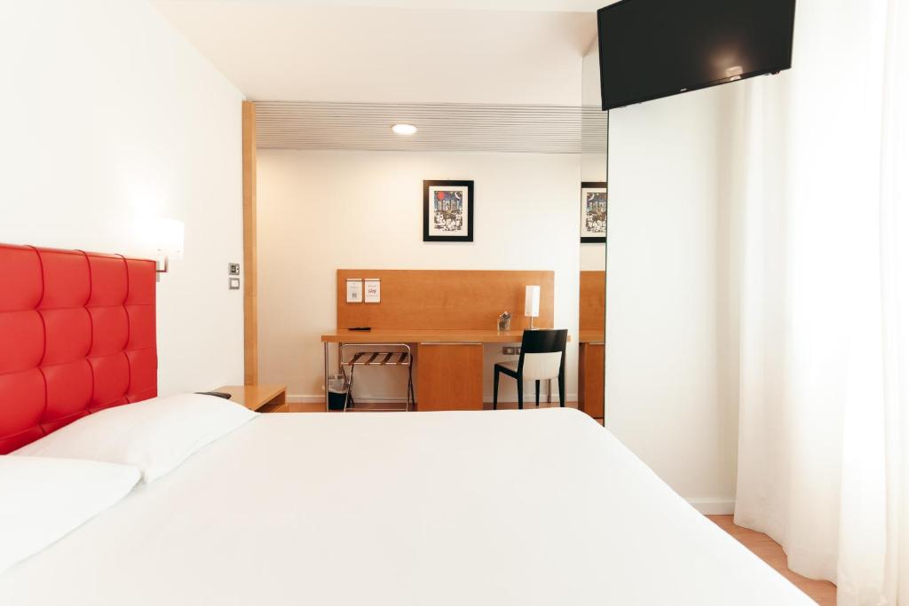 Mūsdienīga viesnīcas numura iekārtojums ar sarkanu gultas galviņu, darba galdu un televizoru. Telpas ir gaišas un gaumīgas, radot patīkamu atmosfēru.