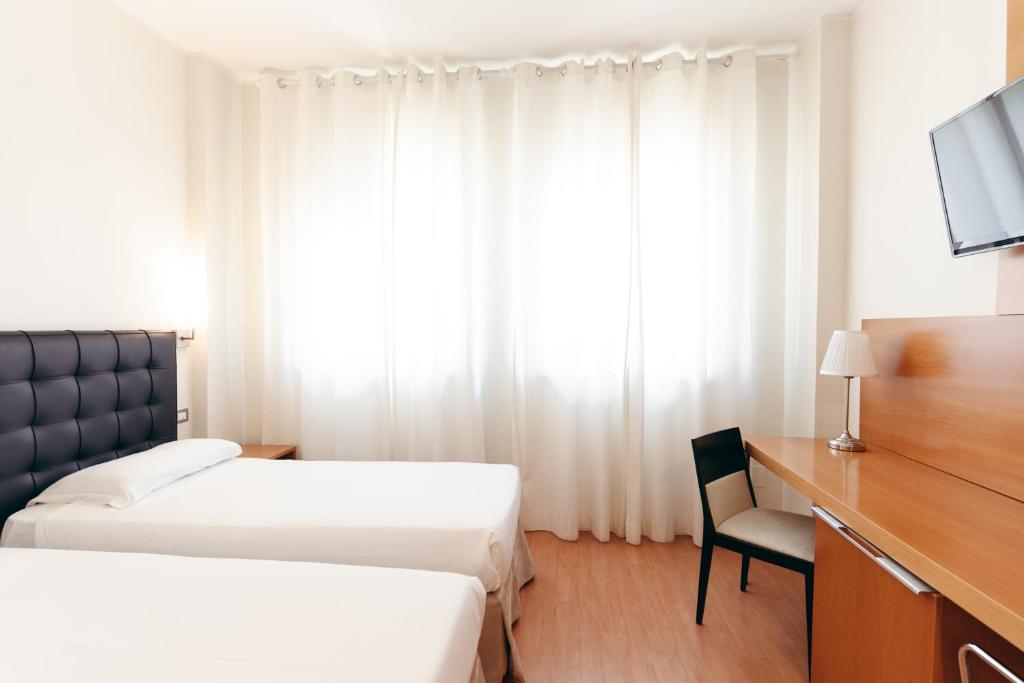 Gaiša viesnīcas numuriņa iekārtojums ar divām atsevišķām gultām, koka rakstāmgaldu un melnu krēslu, kas papildināts ar gaišām aizkariem un televizoru.